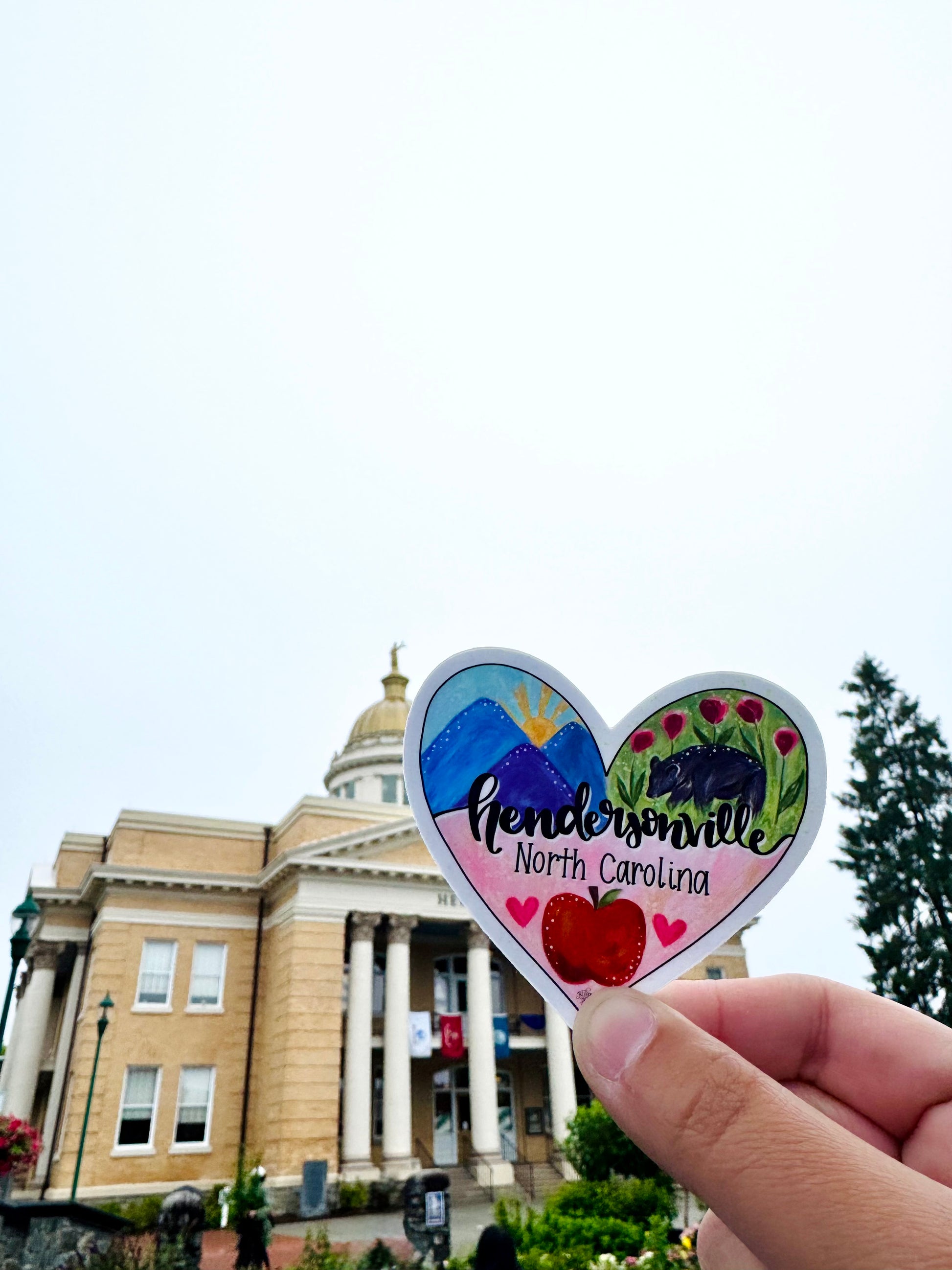 Heart-shaped sticker with 'Hendersonville, North Carolina' text held in front of the Henderson County Historic Courthouse. 