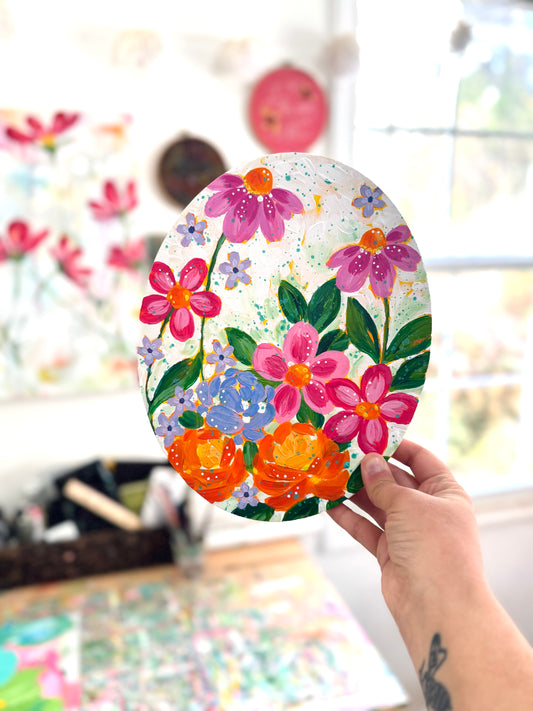 A colorful floral acrylic painting on an oval-shaped canvas with a white background, featuring pink, yellow, and orange flowers with green leaves and yellow underlayers showing through.