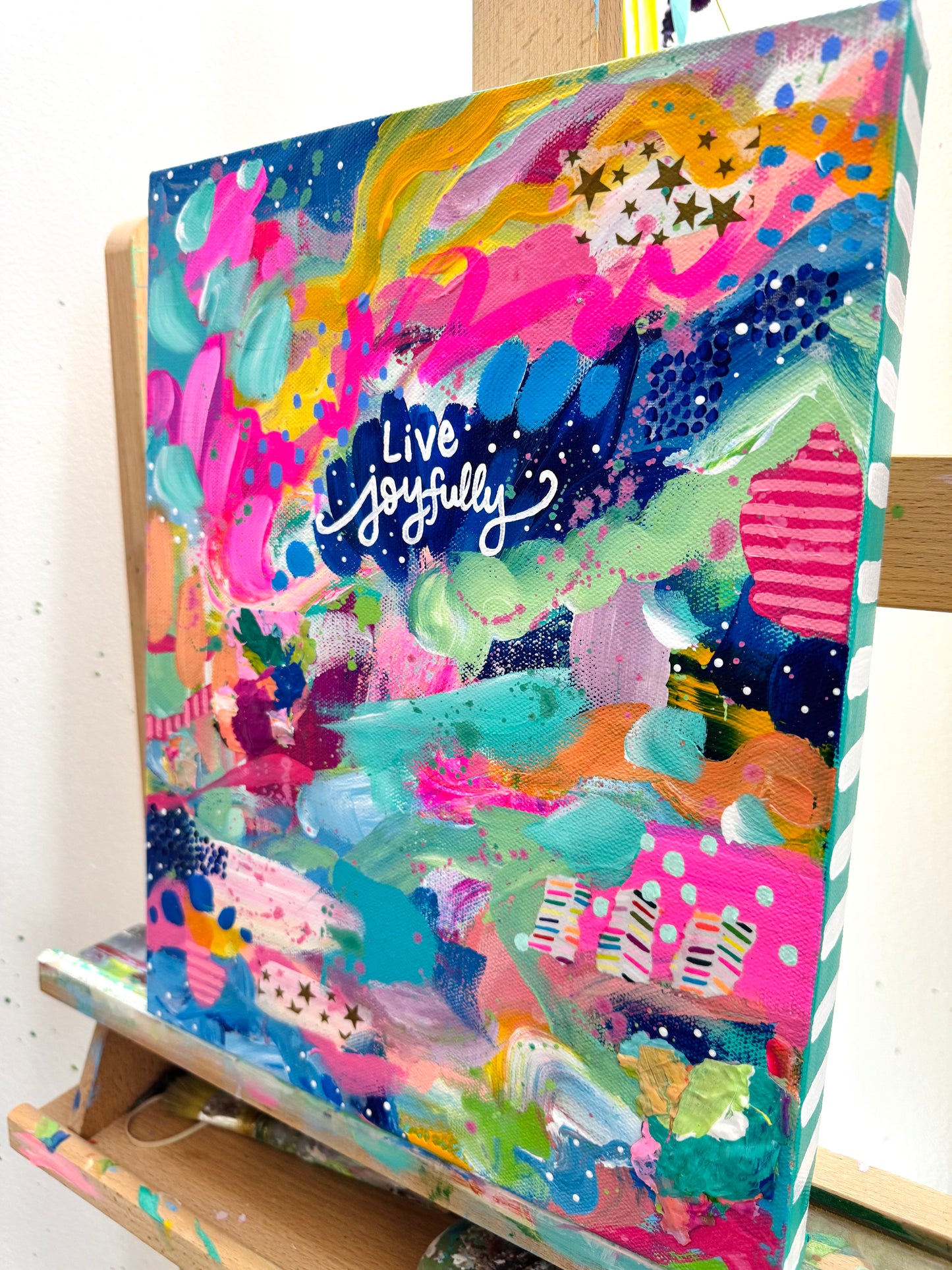 Colorful abstract painting with 'Live Joyfully' text on an easel.