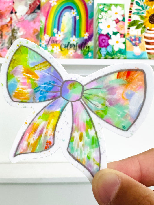 Colorful bow-shaped sticker held by a hand with a blurred background of similar stickers.