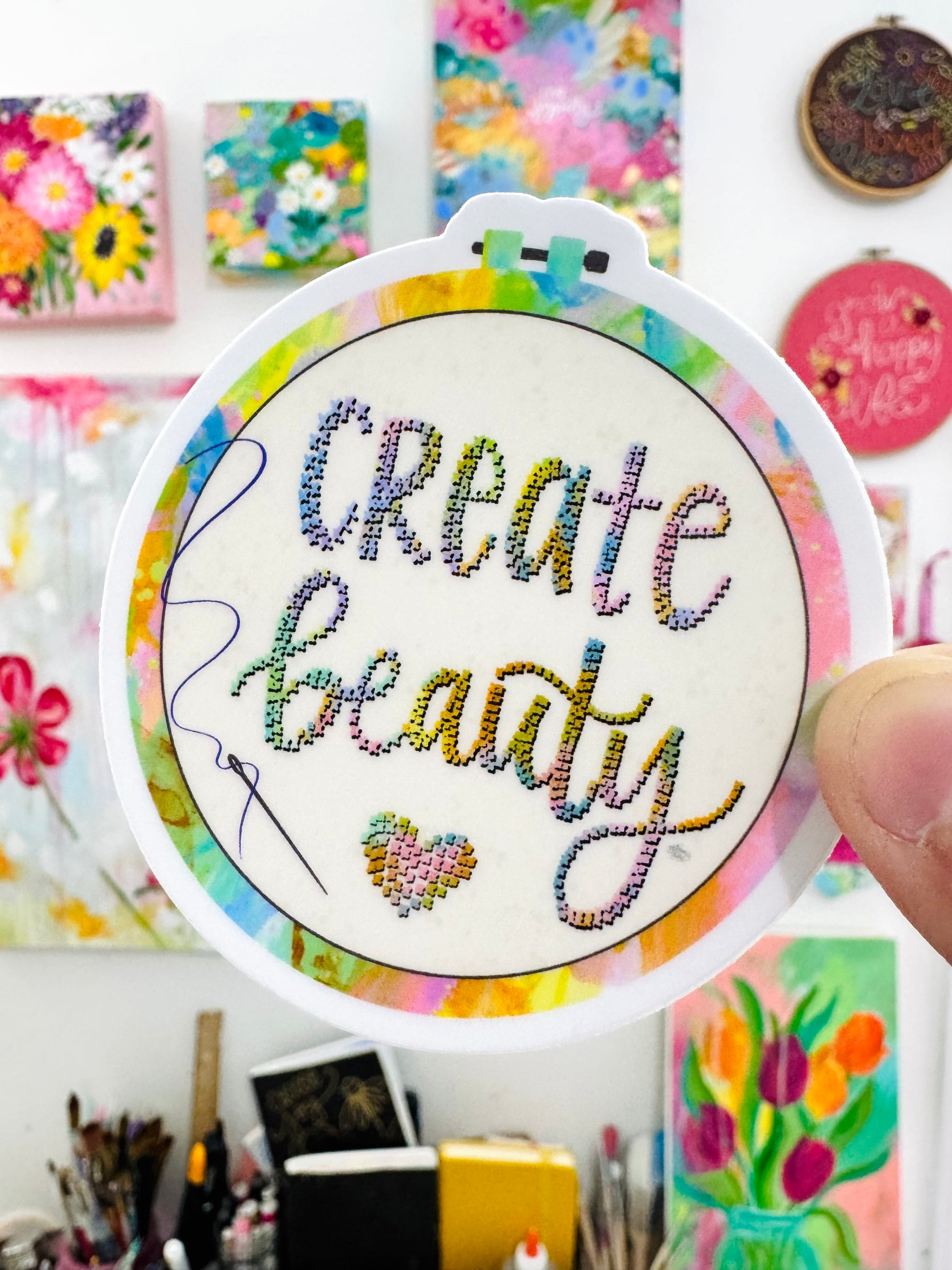 Colorful embroidered patch sticker with 'Create Beauty' text held in front of a colorful background.
