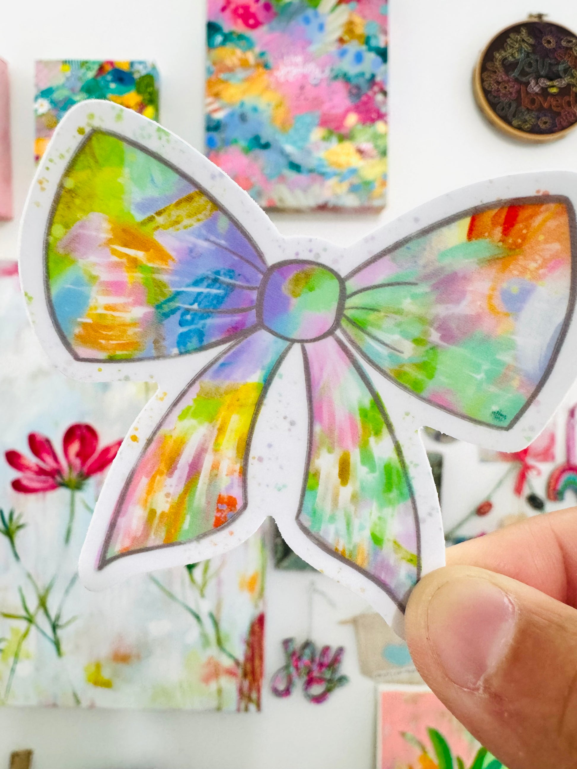 Colorful watercolor bow design held by a hand with floral and abstract art in the background.