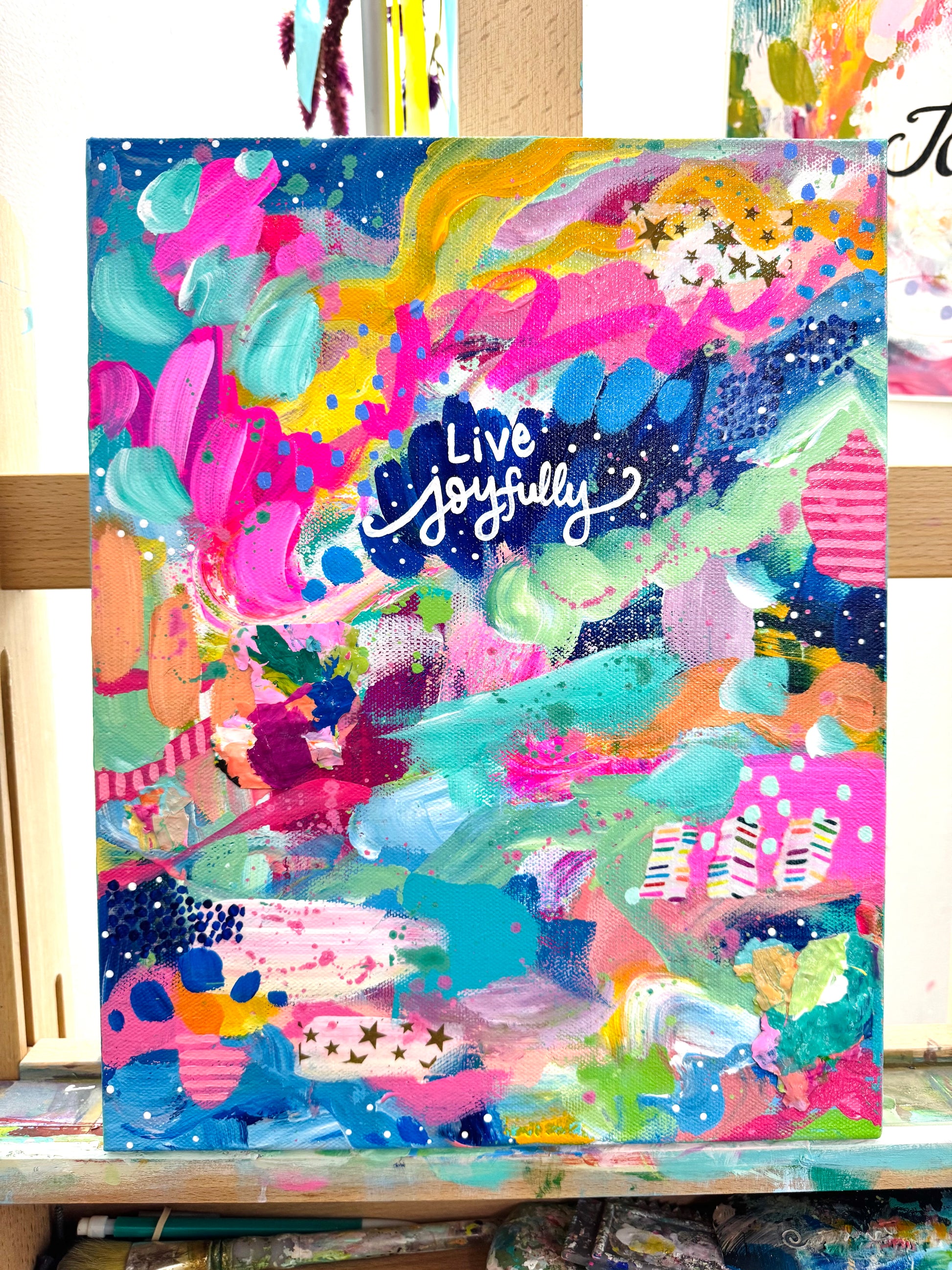 Colorful abstract painting with 'Live Joyfully' text on a wooden surface.