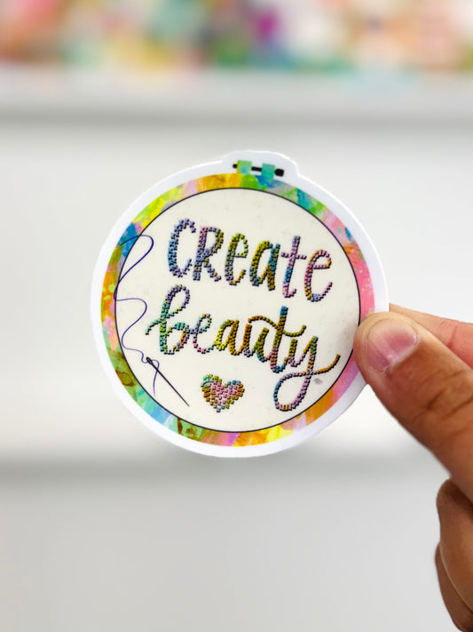 Circular embroidered patch sticker with 'Create beauty' text held by a hand on a light background