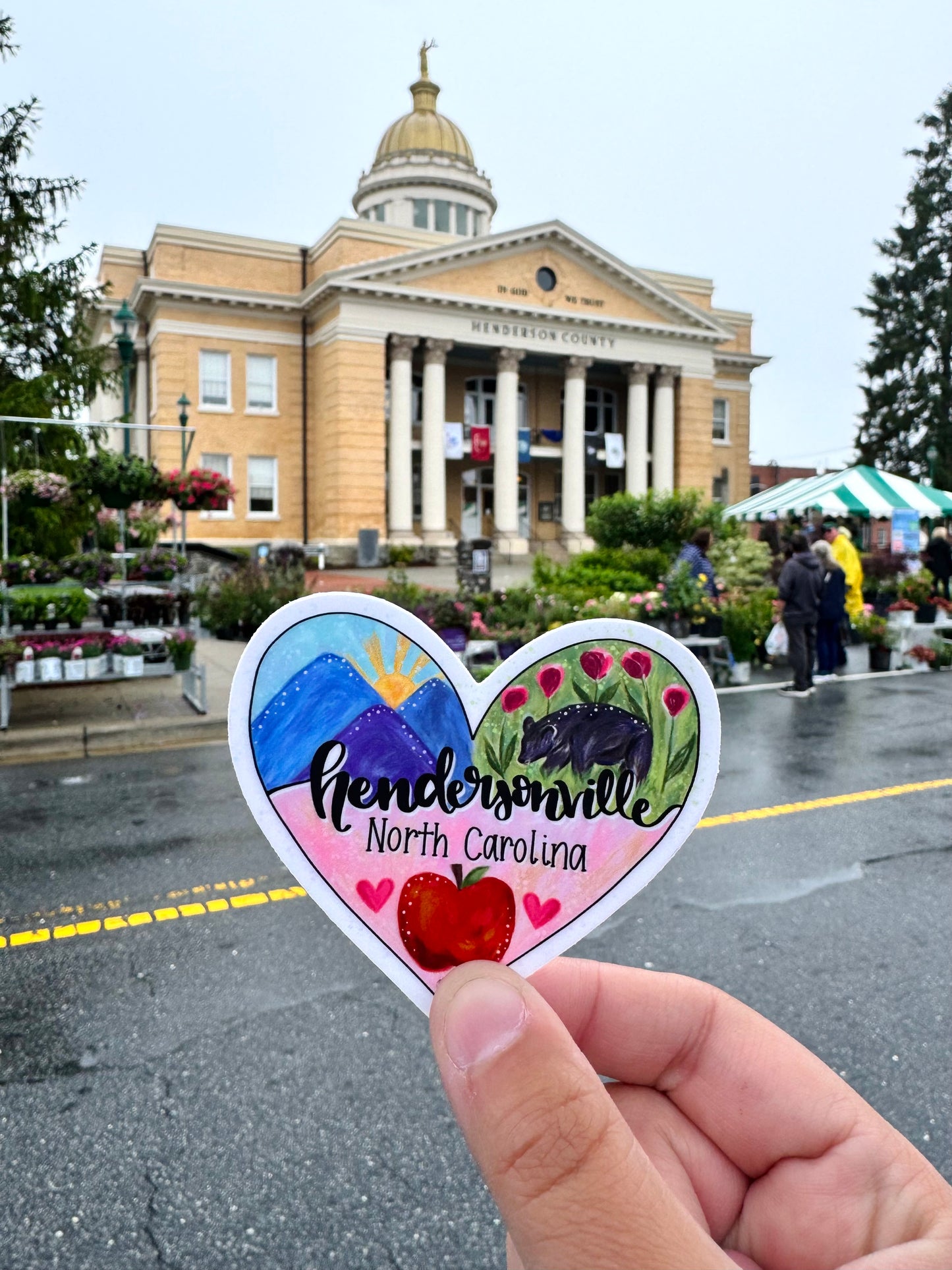 Heart-shaped sticker with 'Hendersonville, North Carolina' text held in front of the Henderson County Historic Courthouse. 