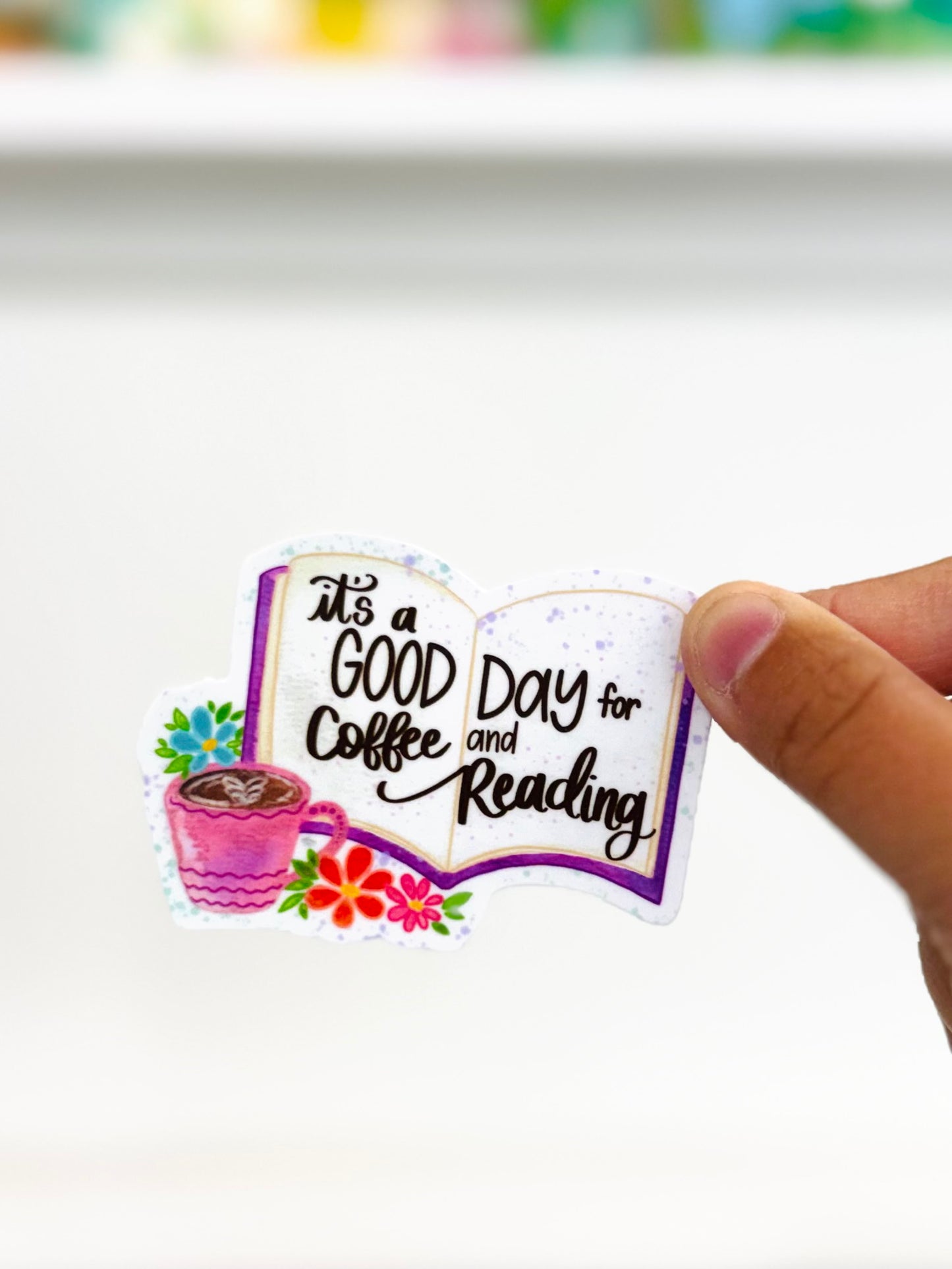 Sticker with 'It's a good day for coffee and reading' text held by a hand on a white background
